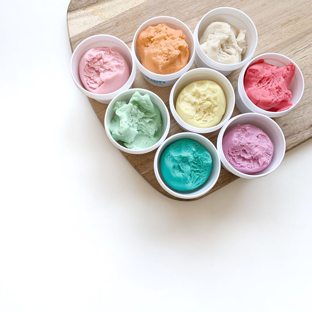 8 tubs of multicoloured dough in tubs sitting on wooden cutting board on a white table